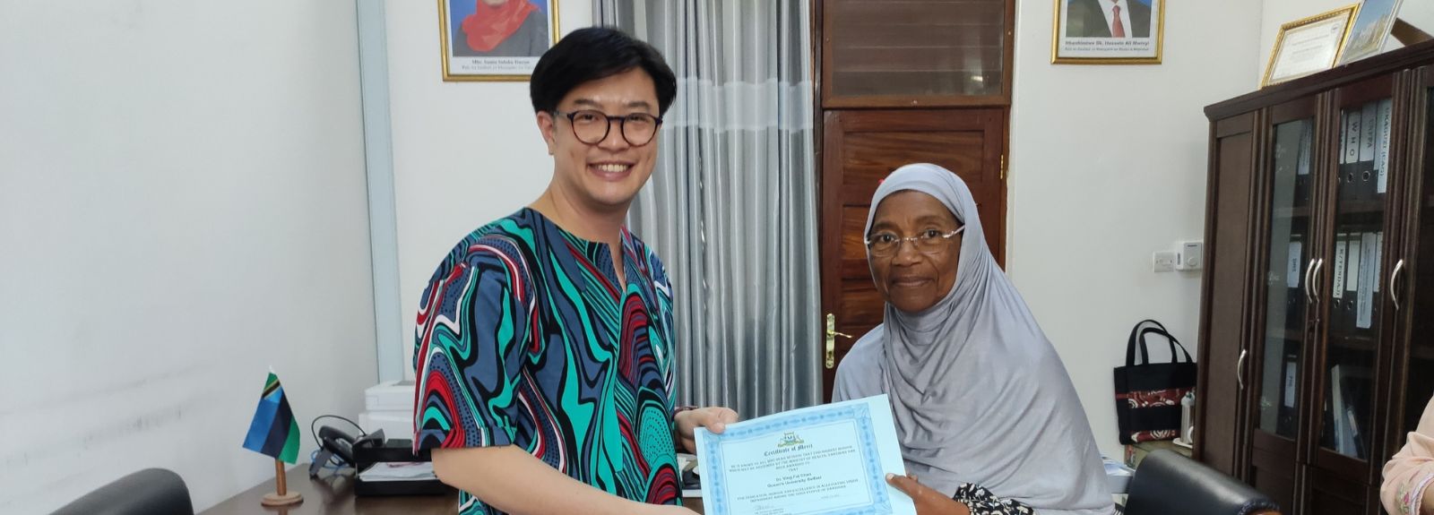 A tall dark haired male wearing glasses and an older head shawl covered women with glasses on, standing in an office, holding a certificate together and smiling at the camera.